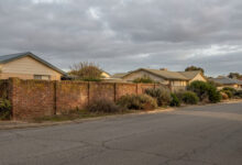 Vue d'une rue résidentielle bordée de maisons avec toits à pignon derrière un mur de briques. Ciel couvert et nuageux.