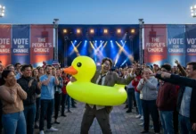 Un homme en costume portant une bouée canard jaune géante sur une scène politique devant une foule enthousiaste.